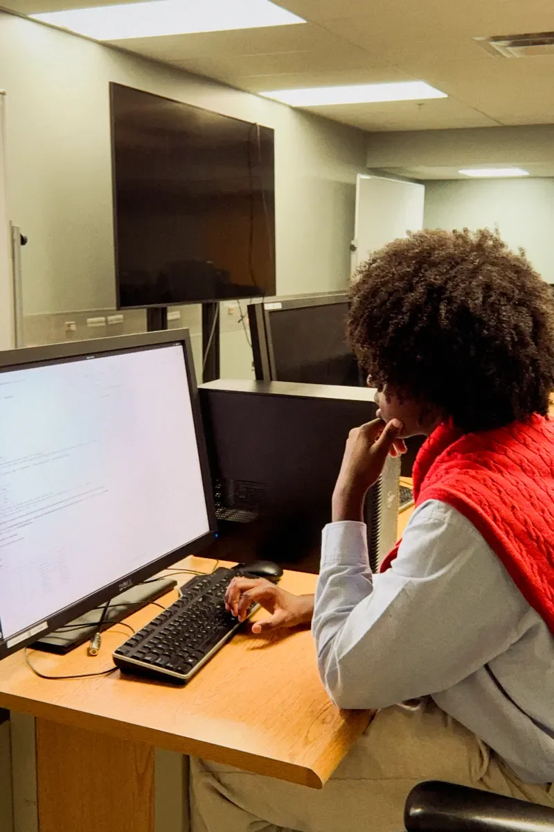 A person typing at a computer lab keyboard with code visible on the monitor