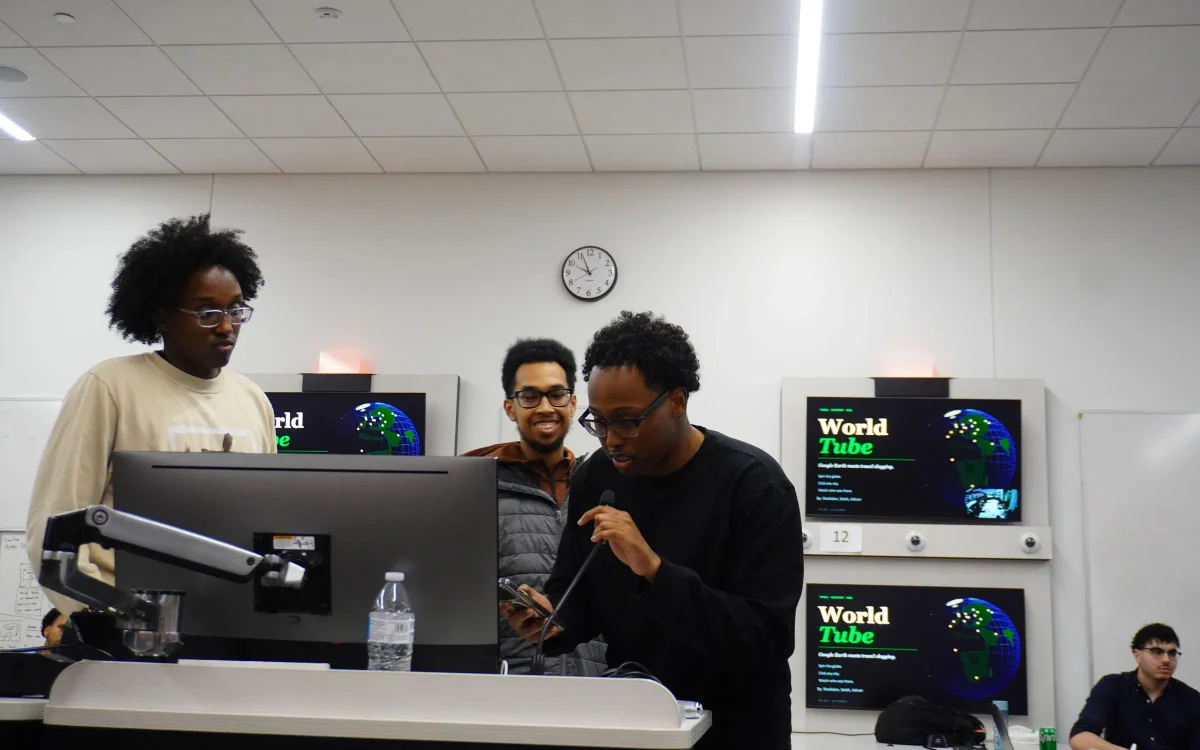 Three students presenting a project slide at a table during the Ideathon, one speaking into a microphone