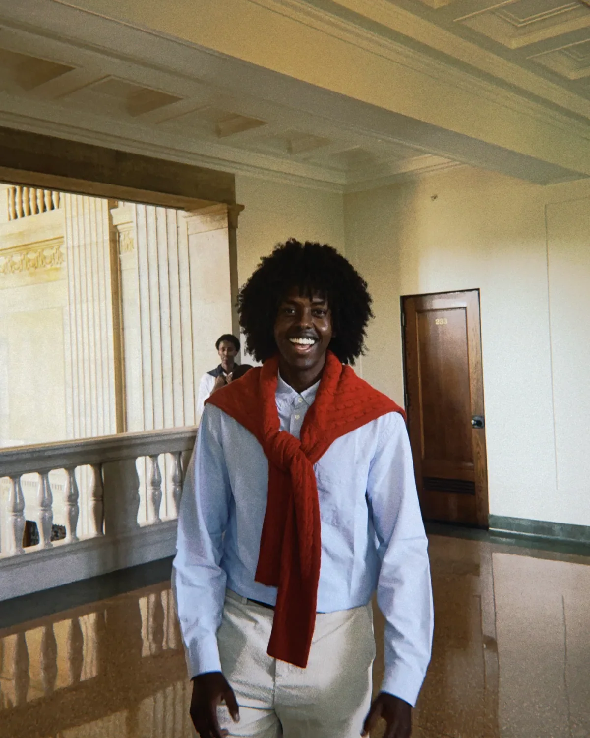 A student smiling in a red scarf, standing in a colonnaded campus hallway